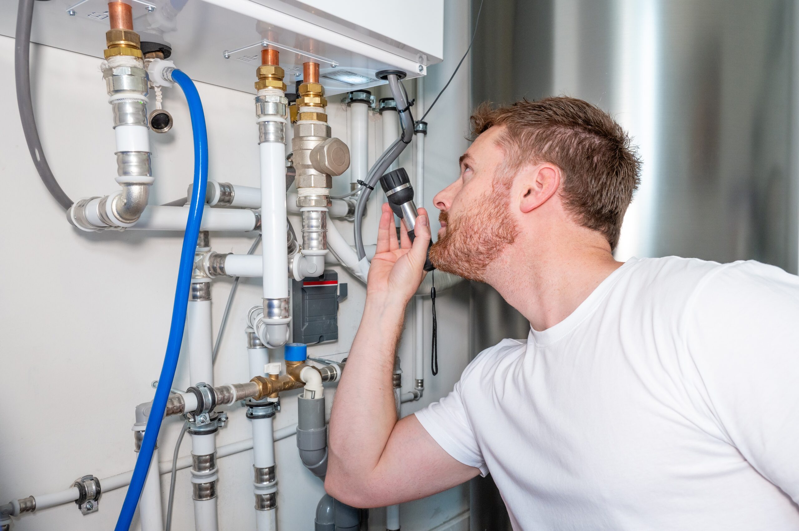 A technician is inspecting a complex network of pipes and fittings connected to a residential or commercial tankless water heater system. He is holding a flashlight and closely examining the connections for signs of leaks, corrosion, or improper installation. The system includes copper, PVC, and PEX piping, along with various valves, sensors, and fittings.