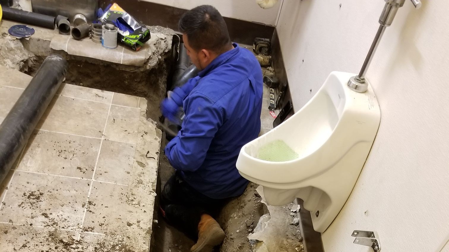 A plumber is working inside a commercial restroom with the floor partially excavated. The technician, dressed in a blue work shirt, is kneeling in the exposed trench, handling piping beneath the tiled floor. A urinal is mounted on the wall nearby, and plumbing tools and supplies are visible on a ledge. The scene captures in-progress pipe repair or replacement, demonstrating hands-on expertise in specialized commercial plumbing services.