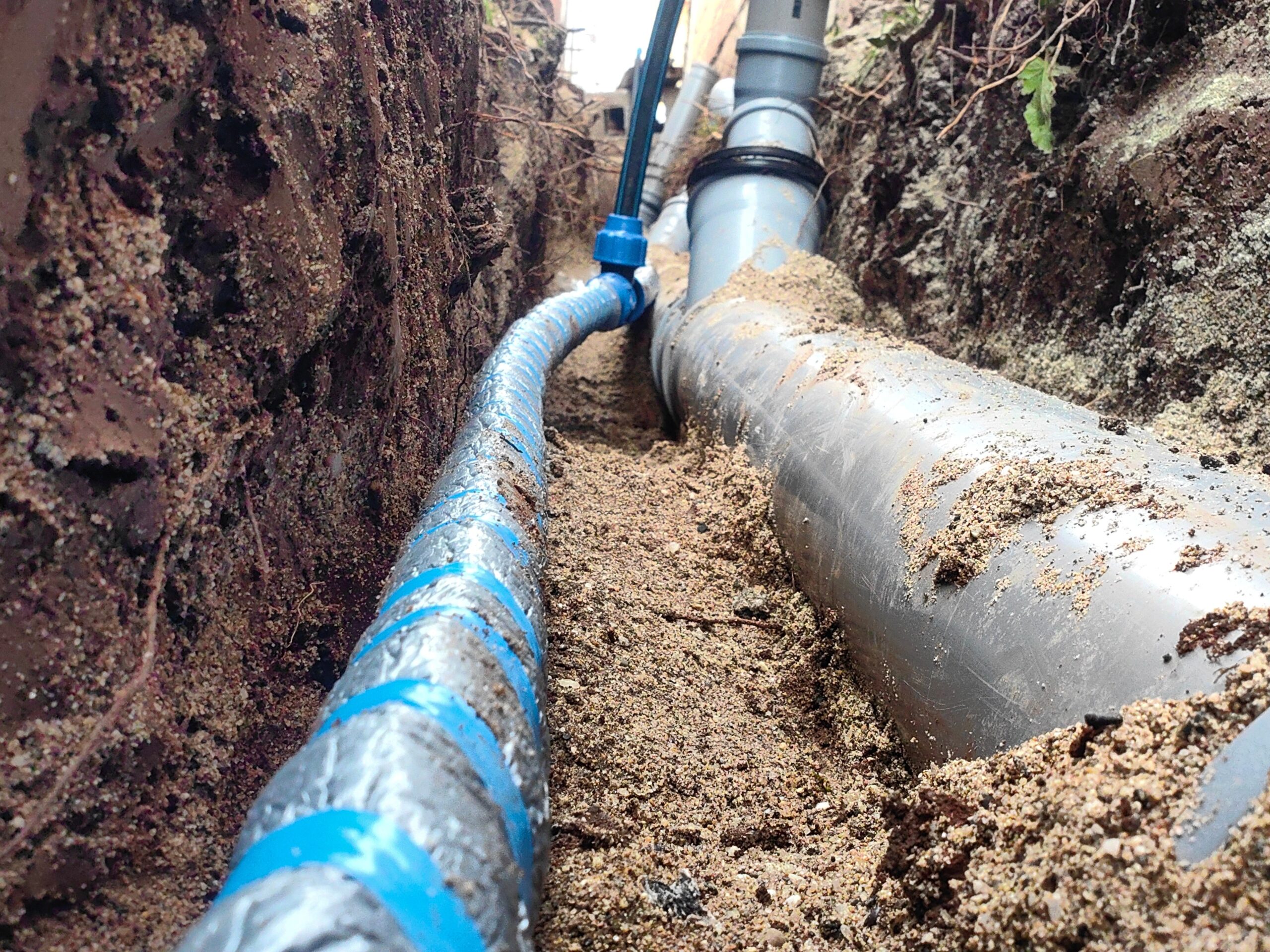Newly installed underground plumbing lines in an open trench. A large gray sewer pipe runs parallel to a smaller blue water supply line, both surrounded by fresh soil and sand bedding. The close-up angle highlights the quality of the installation and the careful alignment of the pipes – ideal for illustrating professional sewer line replacement, water line installation, or other underground plumbing services.