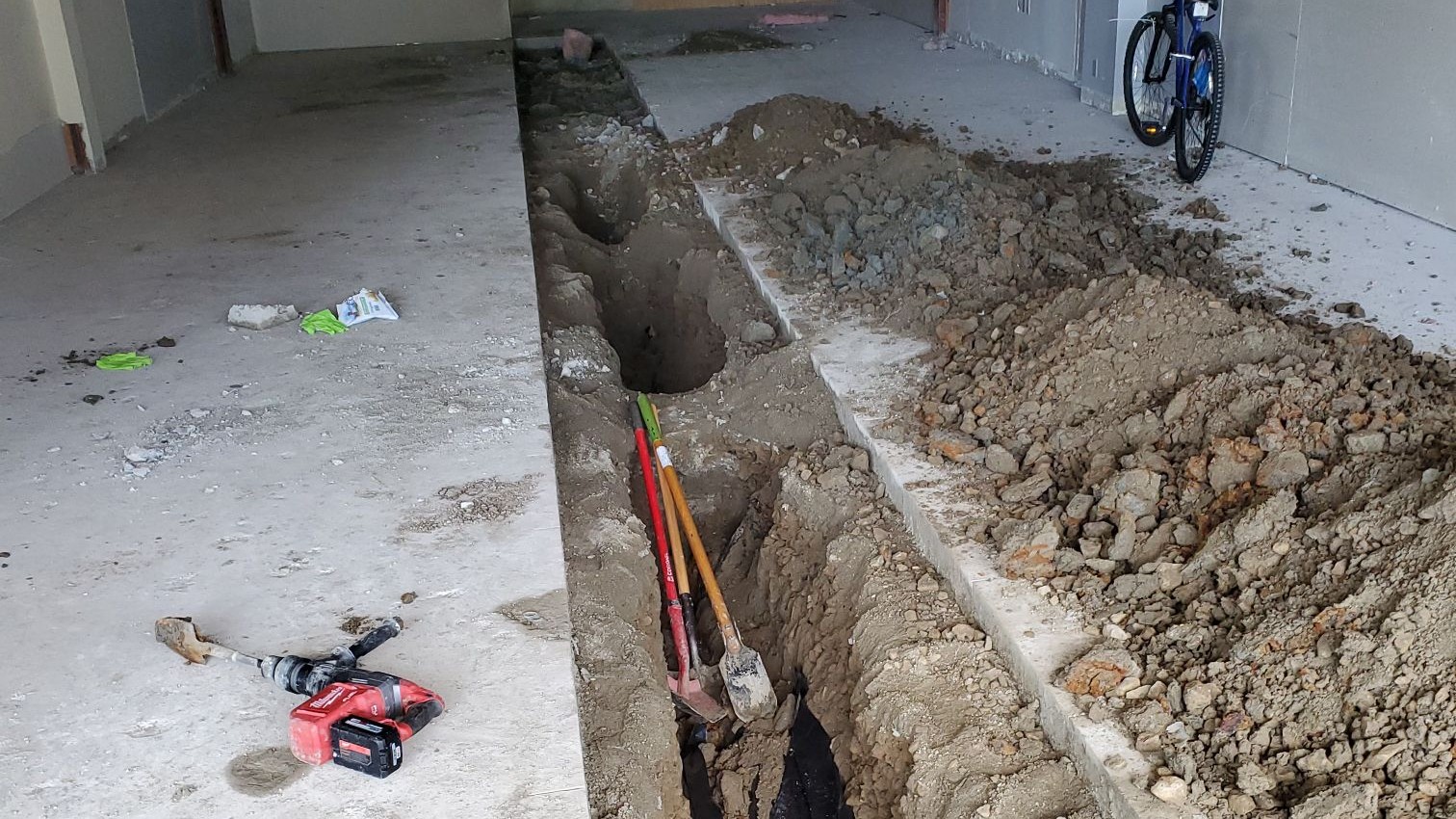 An indoor construction site where a trench has been cut into the concrete floor for plumbing installation or repair. Piles of excavated dirt line one side of the trench, while shovels rest inside the open channel. A red power tool lies on the floor nearby, and a bicycle is visible against the far wall. The scene highlights active specialized plumbing work, such as pipe restoration or new line installation.