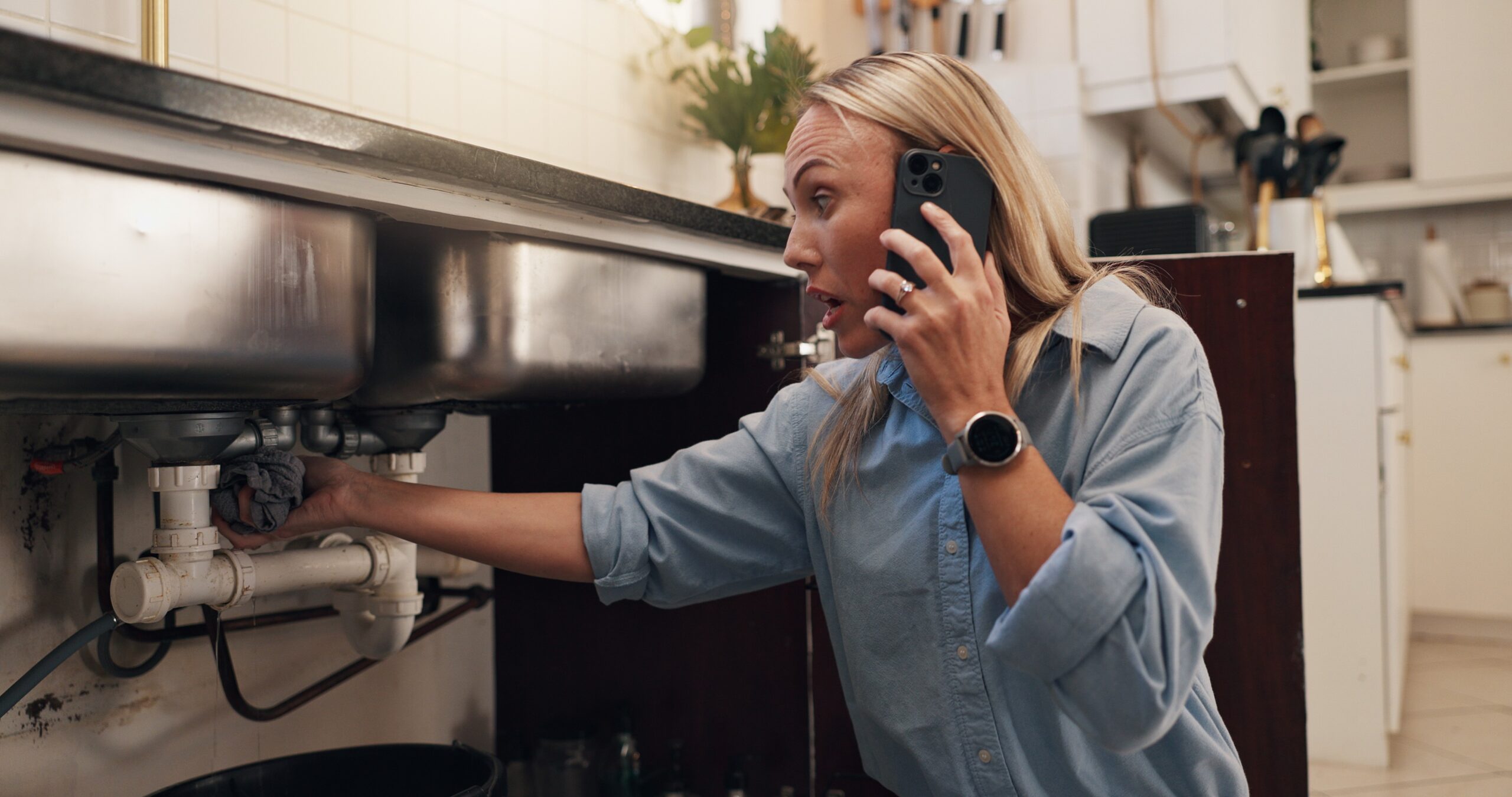 A concerned woman is on the phone while inspecting a plumbing issue under her kitchen sink. She's crouched down, reaching into the cabinet to hold a cloth against a leaking pipe. Her expression shows urgency, likely speaking with a plumber for emergency help.