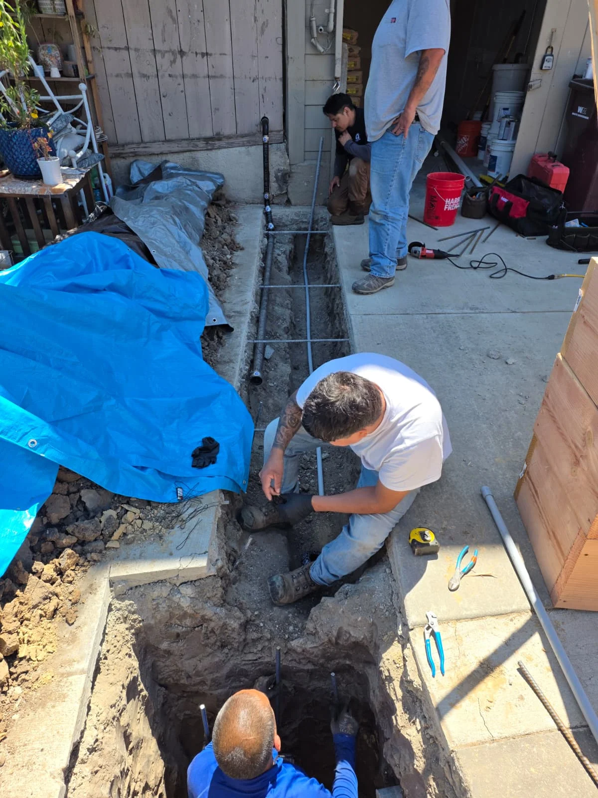 A worker wearing a hard hat and gloves, using a power tool to cut a section of large blue PVC pipe at an outdoor job site. The surrounding dirt and exposed piping indicate an active underground plumbing installation or repair. The worker's focused posture and use of specialized tools highlight precision and expertise in large-scale water or sewer line projects.