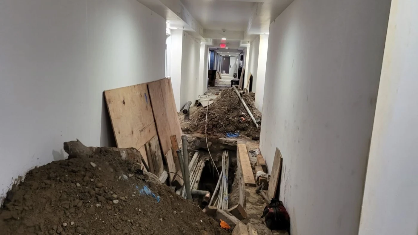A long indoor hallway is undergoing major plumbing work. The floor has been opened up to expose multiple pipes running through a trench, with large piles of dirt and construction materials lining the corridor. Wooden boards and tools are placed along the sides, indicating active repair or replacement of the building’s plumbing infrastructure. This scene illustrates large-scale pipe restoration or excavation in a commercial or multi-unit residential property.