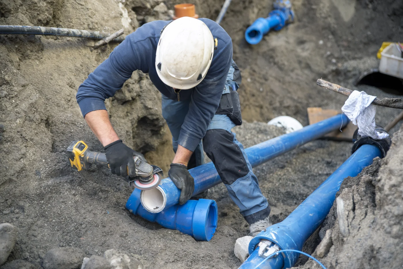 A worker wearing a hard hat and gloves, using a power tool to cut a section of large blue PVC pipe at an outdoor job site. The surrounding dirt and exposed piping indicate an active underground plumbing installation or repair. The worker’s focused posture and use of specialized tools highlight precision and expertise in large-scale water or sewer line projects.