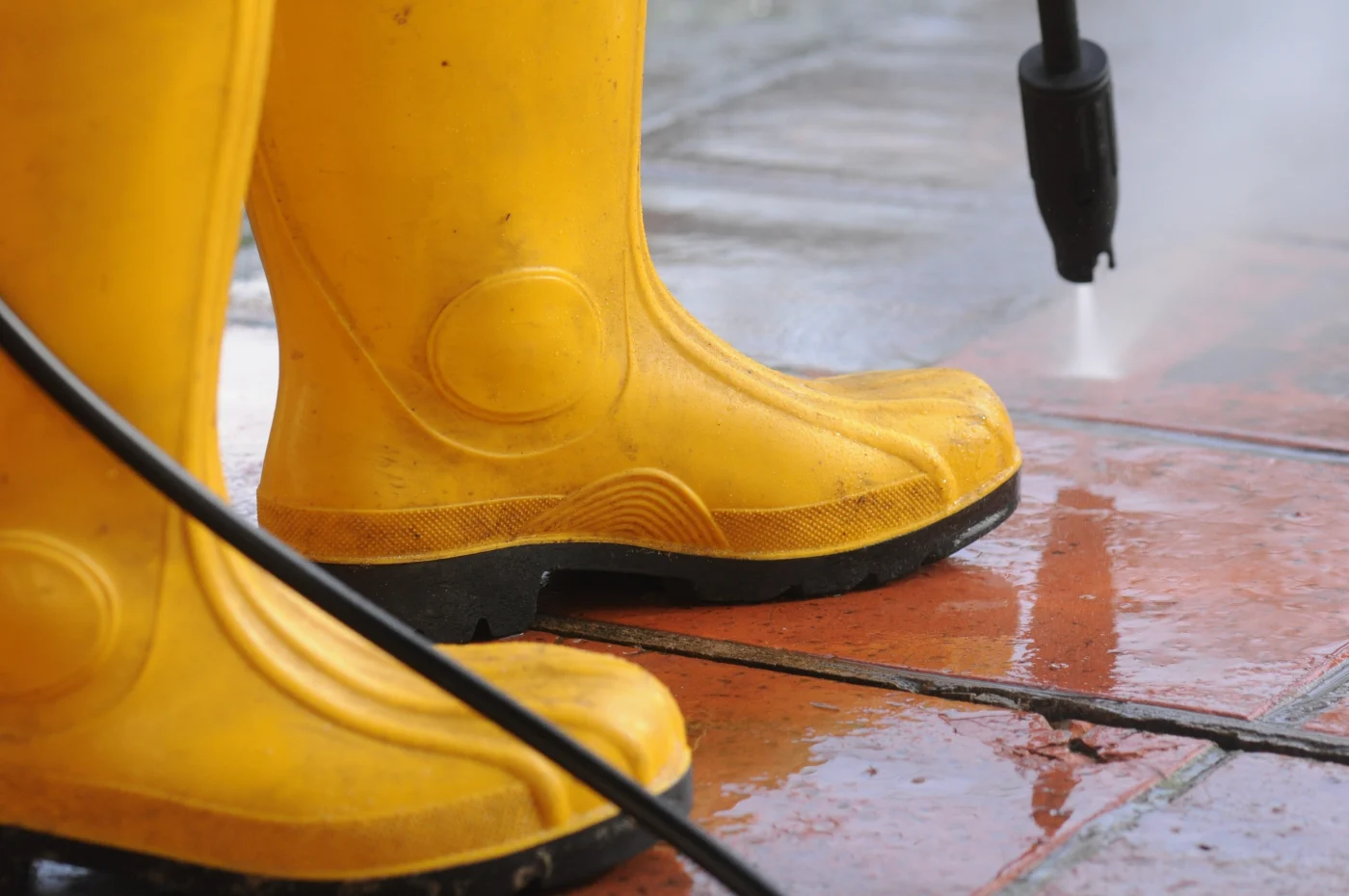 A plumber with yellow galoshes pressure washing a red sidewalk.