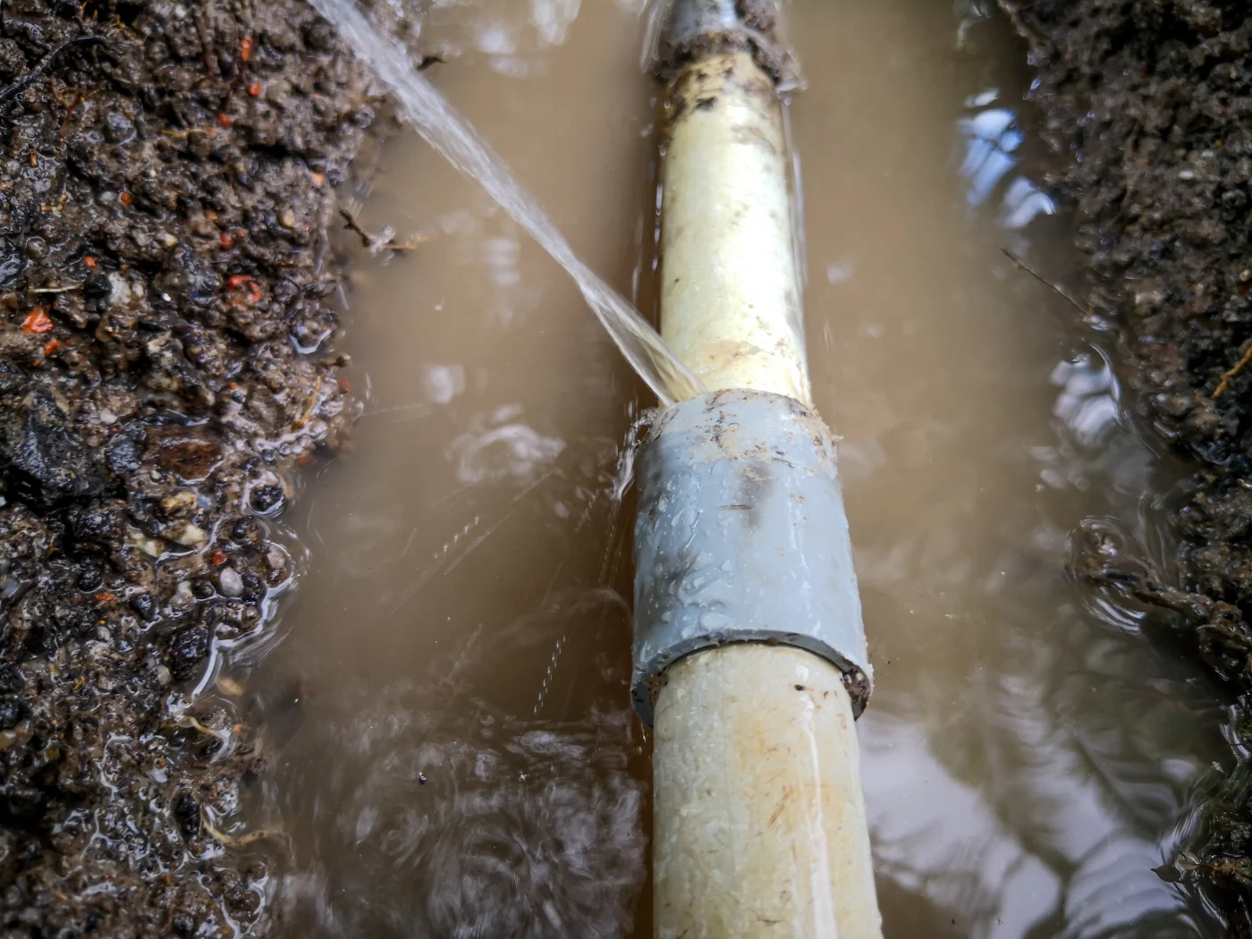 A leaking underground pipe is spraying water into a muddy trench during plumbing and drain repair.