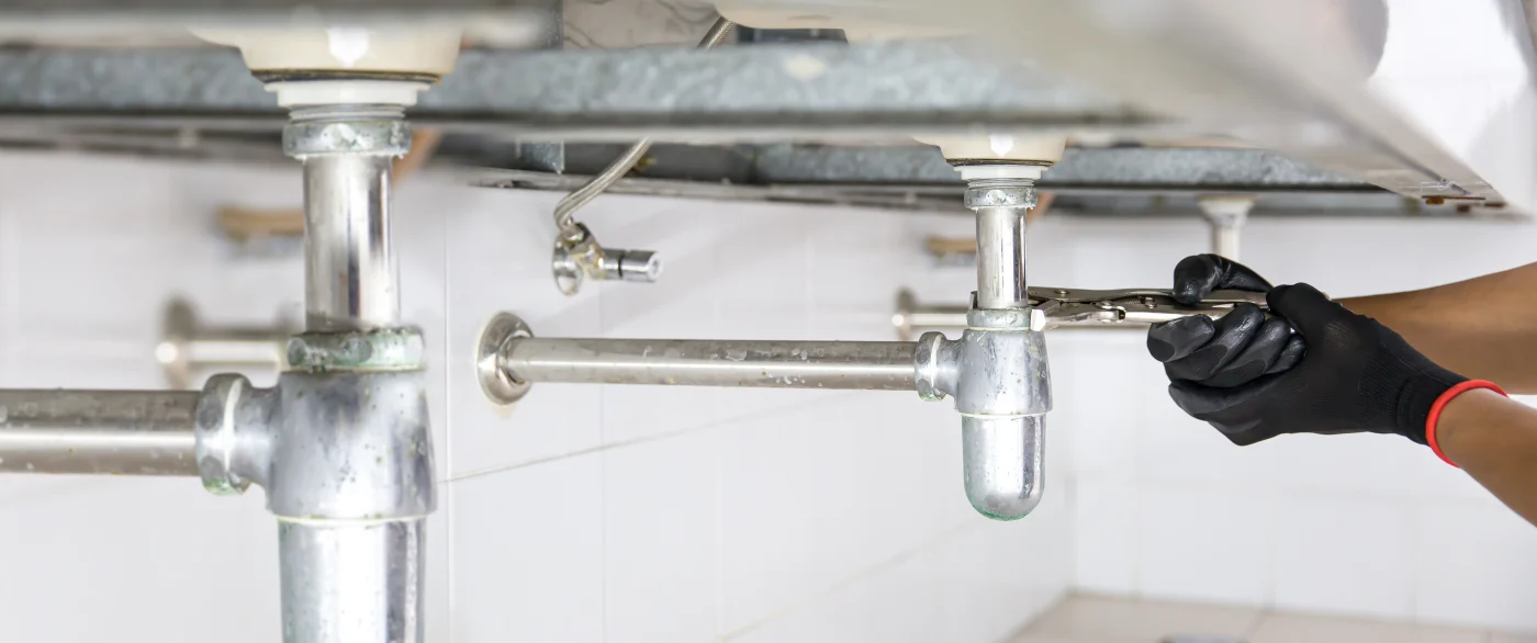 A technician wearing black gloves and using pliers to tighten a metal drain pipe beneath a sink.