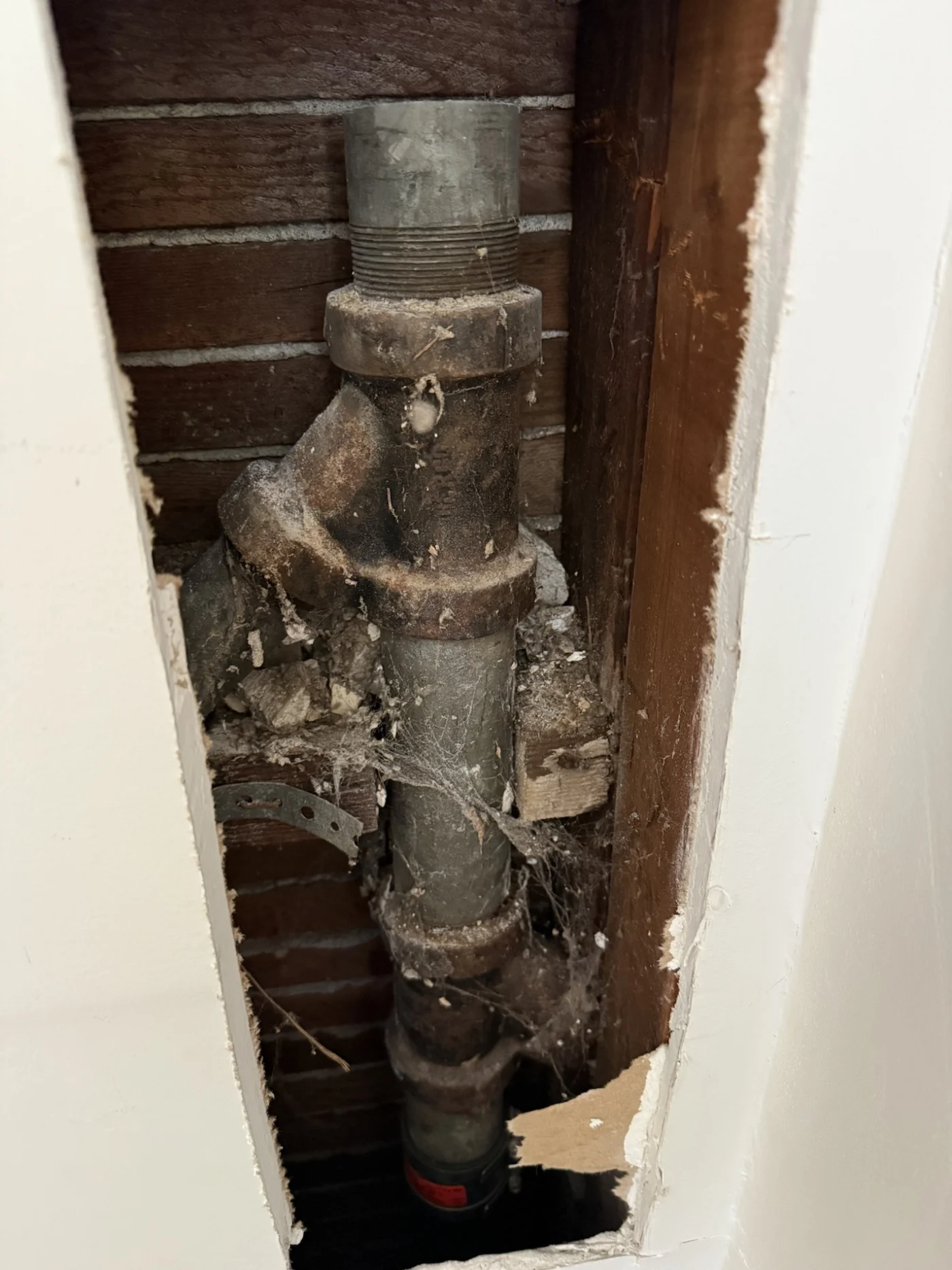 Wall opening exposes an old vertical drain pipe with fittings, debris, and framing during plumbing inspection or repair.
