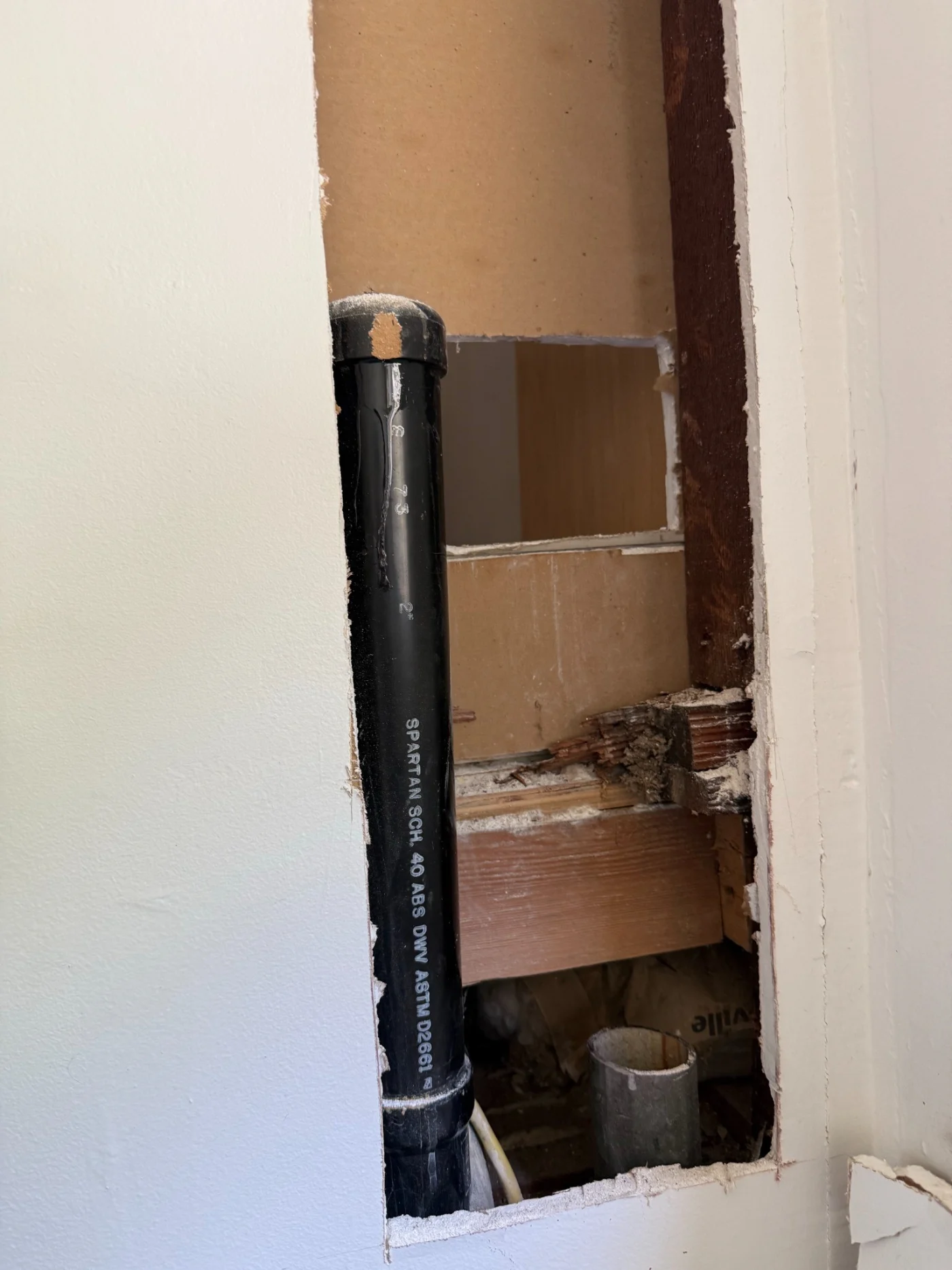 Wall opening exposes black drain pipe, wood framing, and cut drywall during plumbing and drain repair.