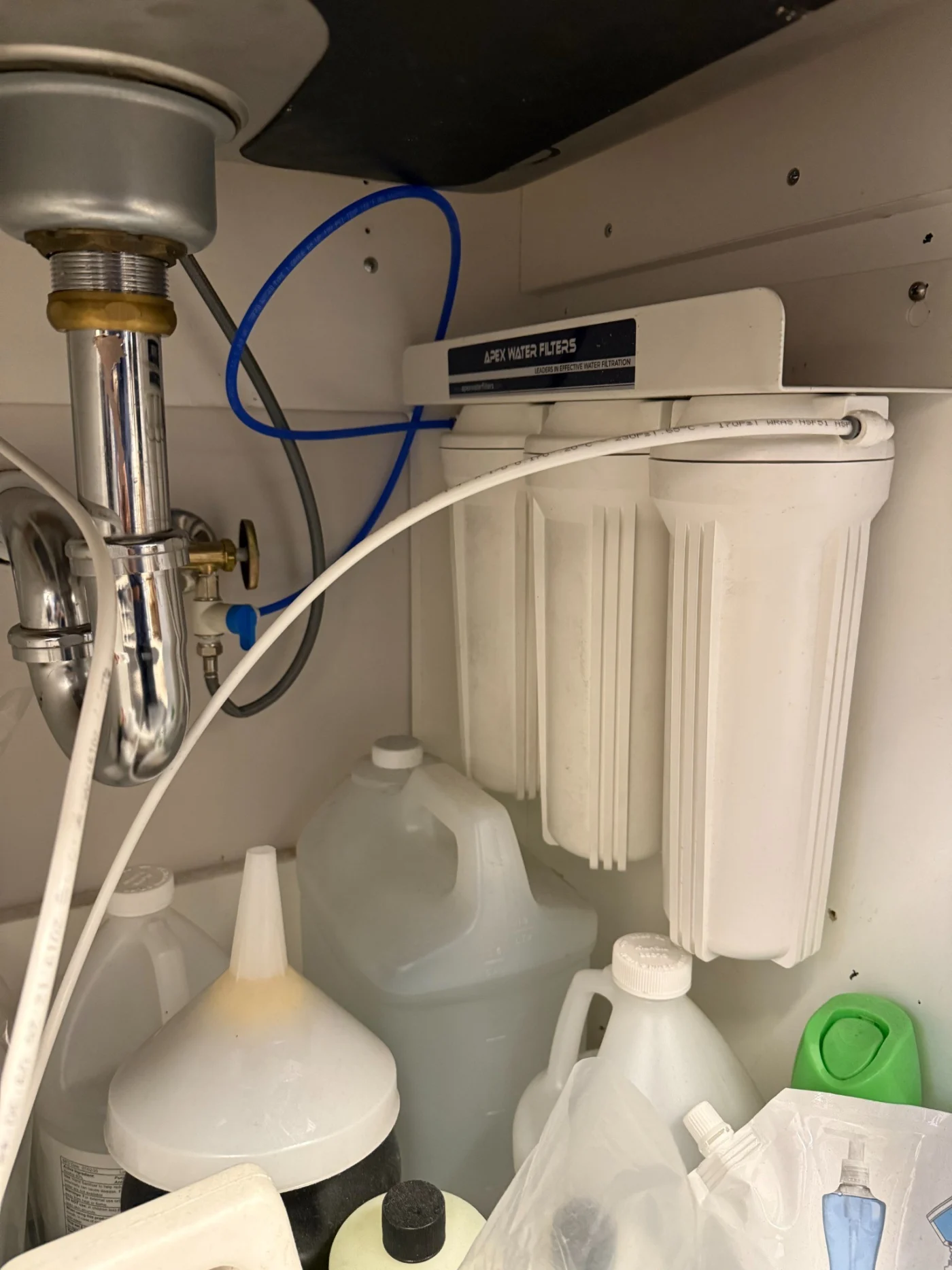 Under-sink water filtration system with chrome drain trap, tubing, and stored cleaning supplies inside a cabinet.