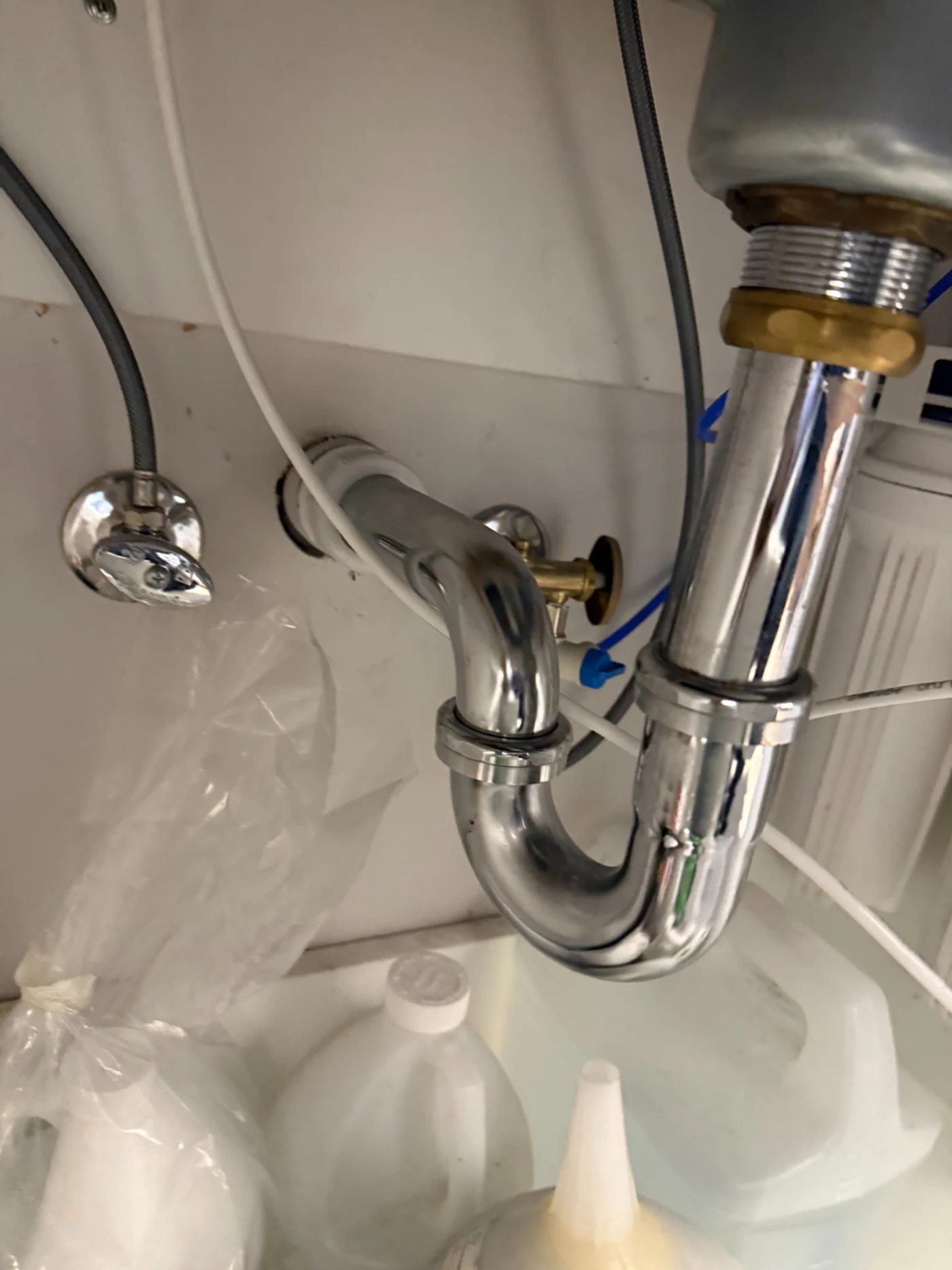 Under-sink chrome drain trap with water supply lines and household cleaning containers visible inside a cabinet.