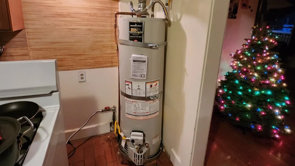A traditional gas water heater is installed beside a kitchen area in a home with visible copper piping and vent connections.