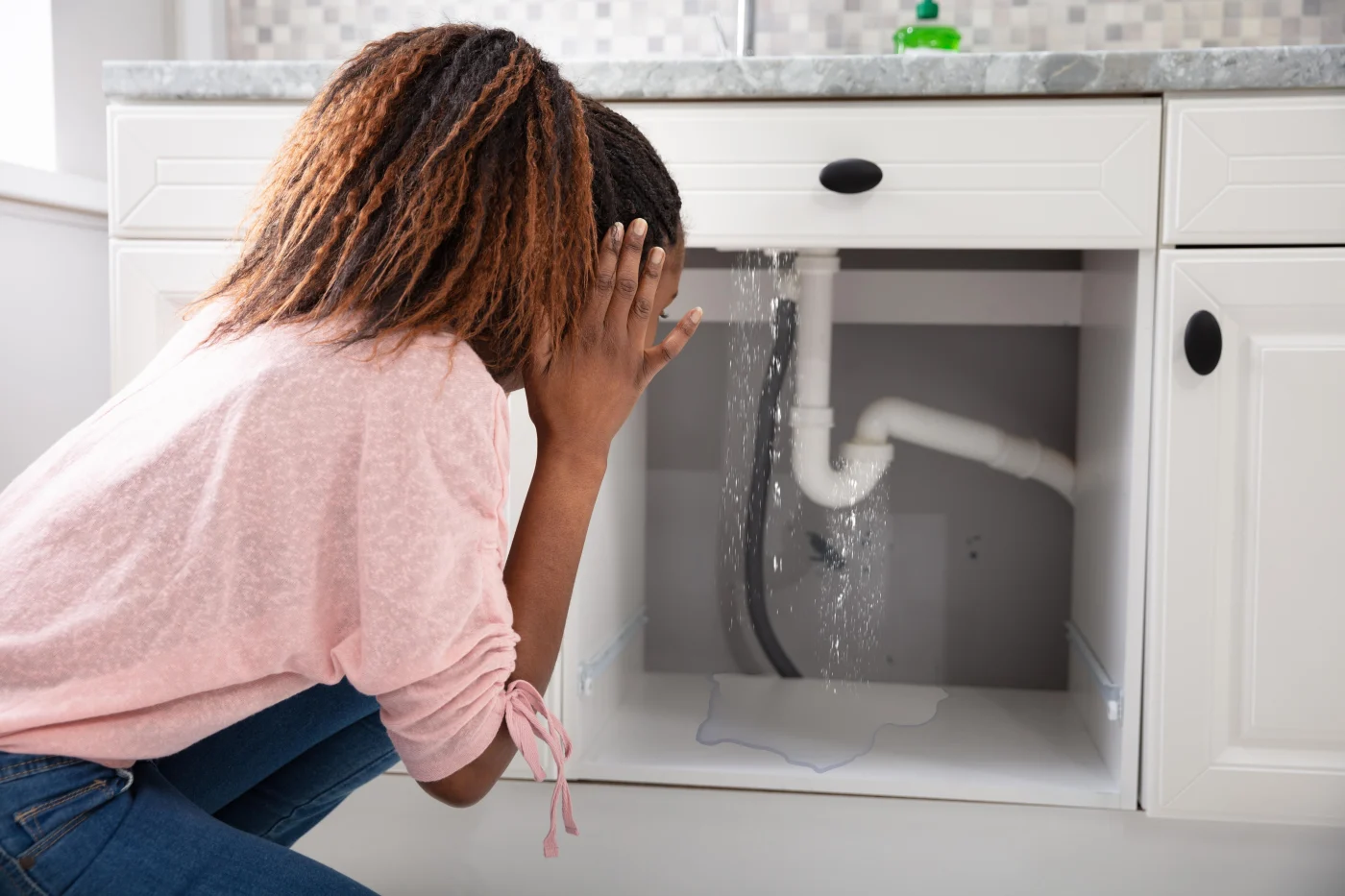 A woman reacts to water leaking from a sink pipe under a bathroom vanity, with water pooling on the cabinet floor.