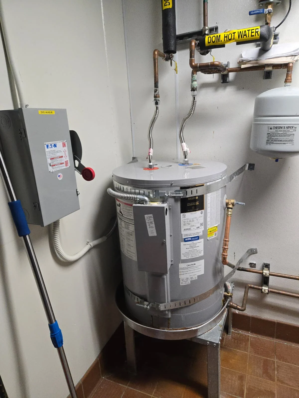 Commercial water heater with copper piping, labeled hot water line, expansion tank, and electrical panel in a utility room.