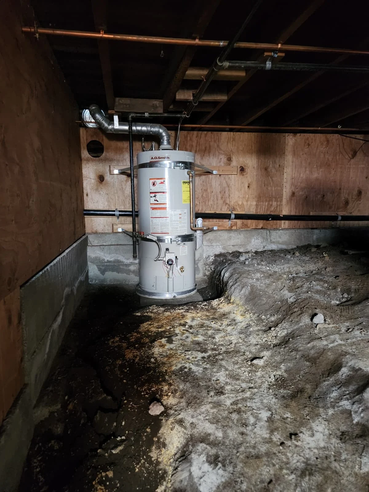 Water heater installed in a crawl space with visible piping, venting, exposed framing, and dirt floor.