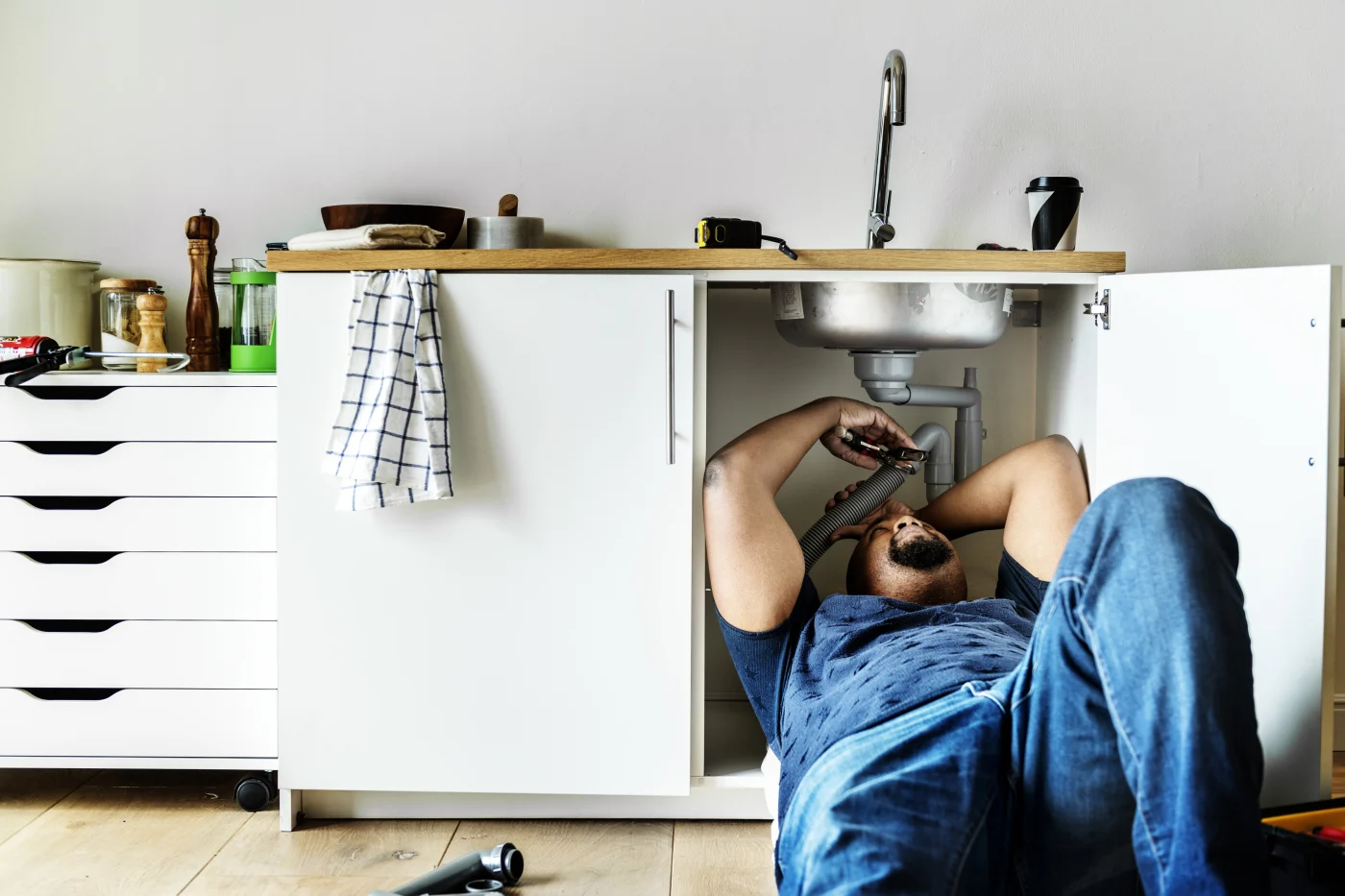 A plumber works under a kitchen sink with the cabinet doors open while repairing the drain pipes.