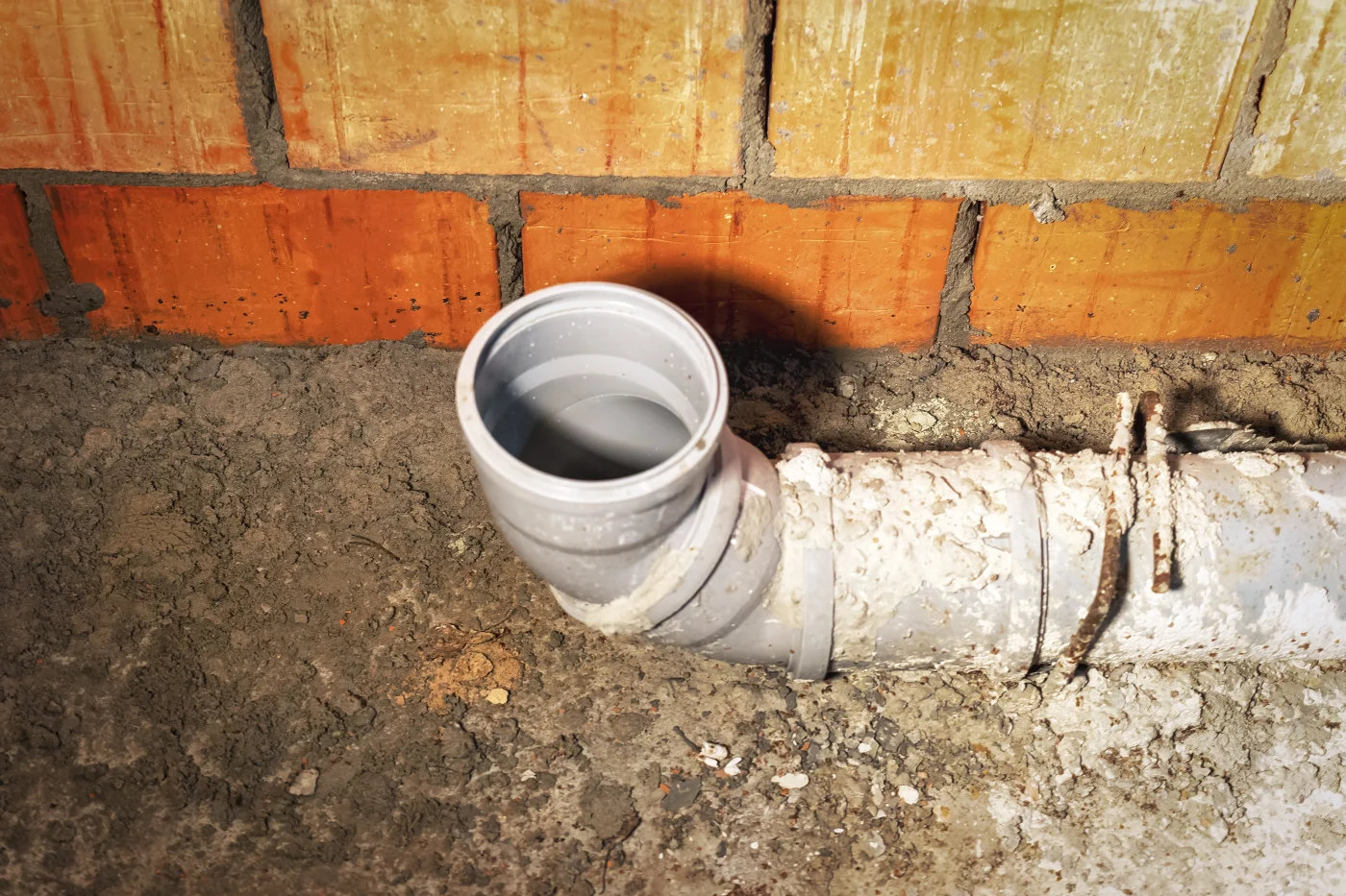 Open gray sewer cleanout pipe beside a brick wall, with dirt, mortar, rough concrete, and old white pipe during sewer replacement.