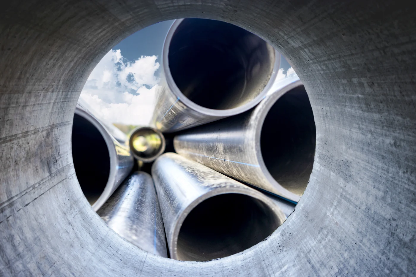 Stacked black sewer replacement pipes viewed through an open pipe end, with circular openings, blue guide stripes, and cloudy sky beyond.