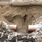 Snowy excavation trench with exposed sewer pipes, dirt walls, safety tape, drainage grate, and construction debris during sewer replacement.