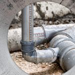 Gray PVC sewer pipes inside a concrete opening with pipe joints, fittings, dirt, debris, and rough construction surface around the plumbing.