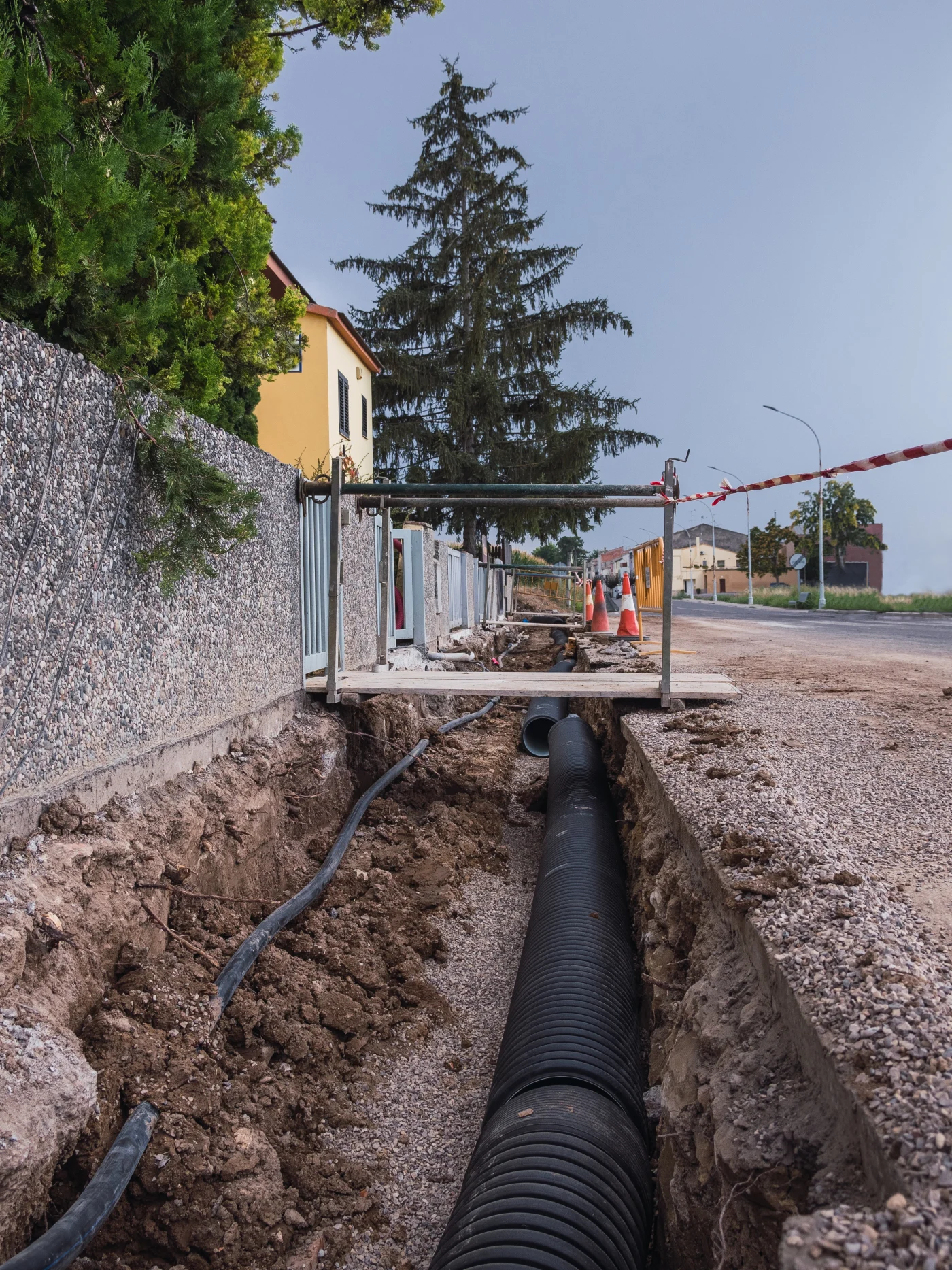Street trench with black corrugated sewer pipe, gravel, dirt walls, safety cones, caution tape, temporary walkway, and nearby homes.