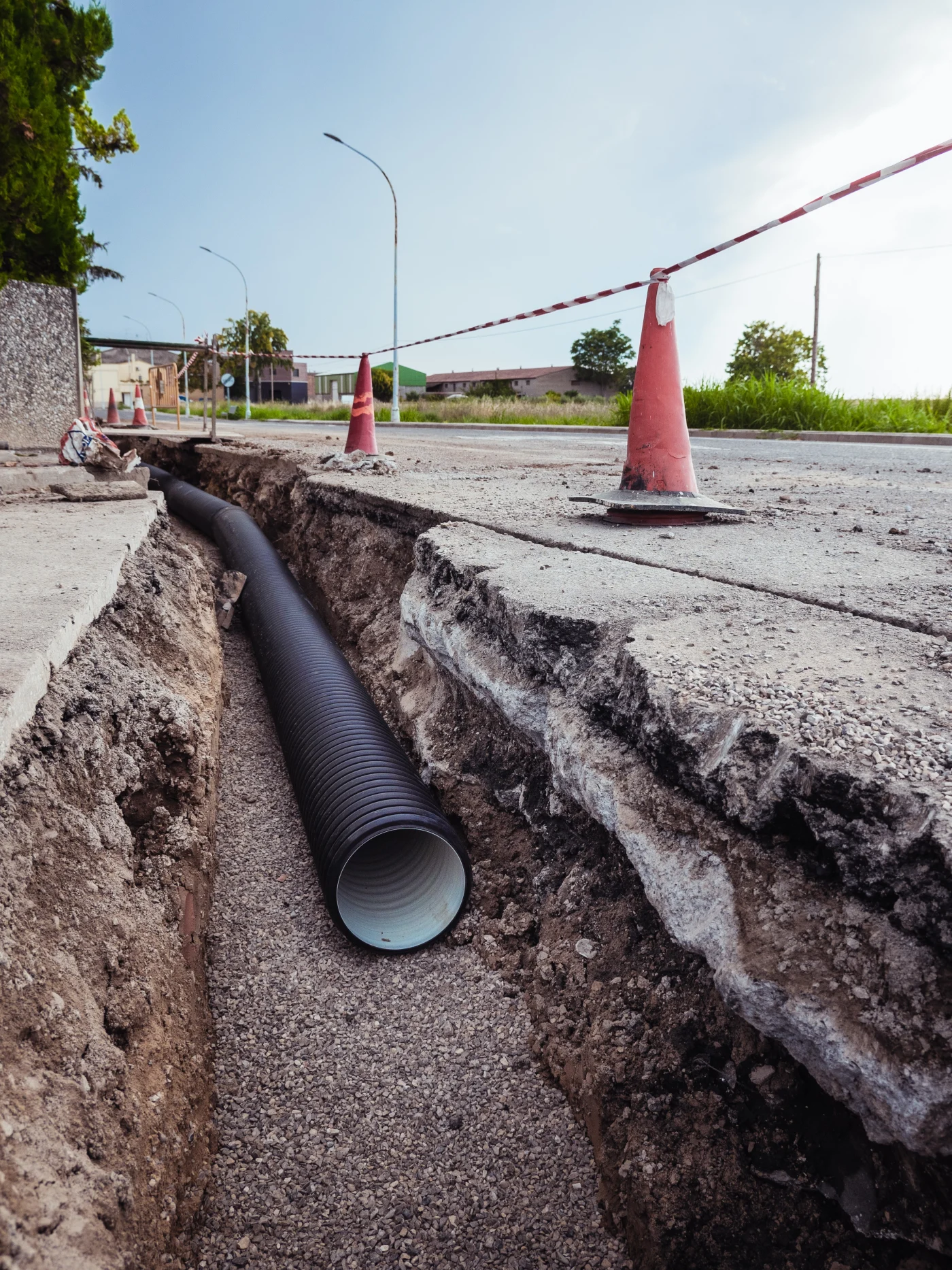 Excavated trench with exposed black sewer pipes, standing water, muddy soil, gravel, and rough pipe opening during sewer replacement