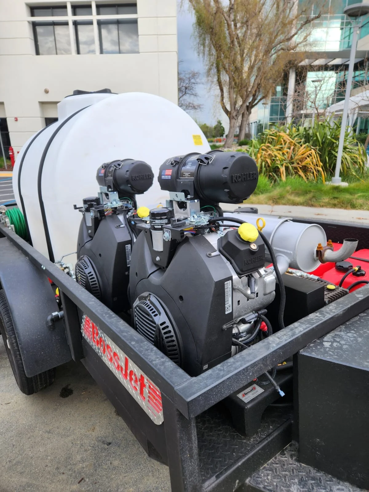 Hydro jetting equipment trailer with dual engines, white water tank, hose reel, pipes, and fittings parked beside a commercial building.