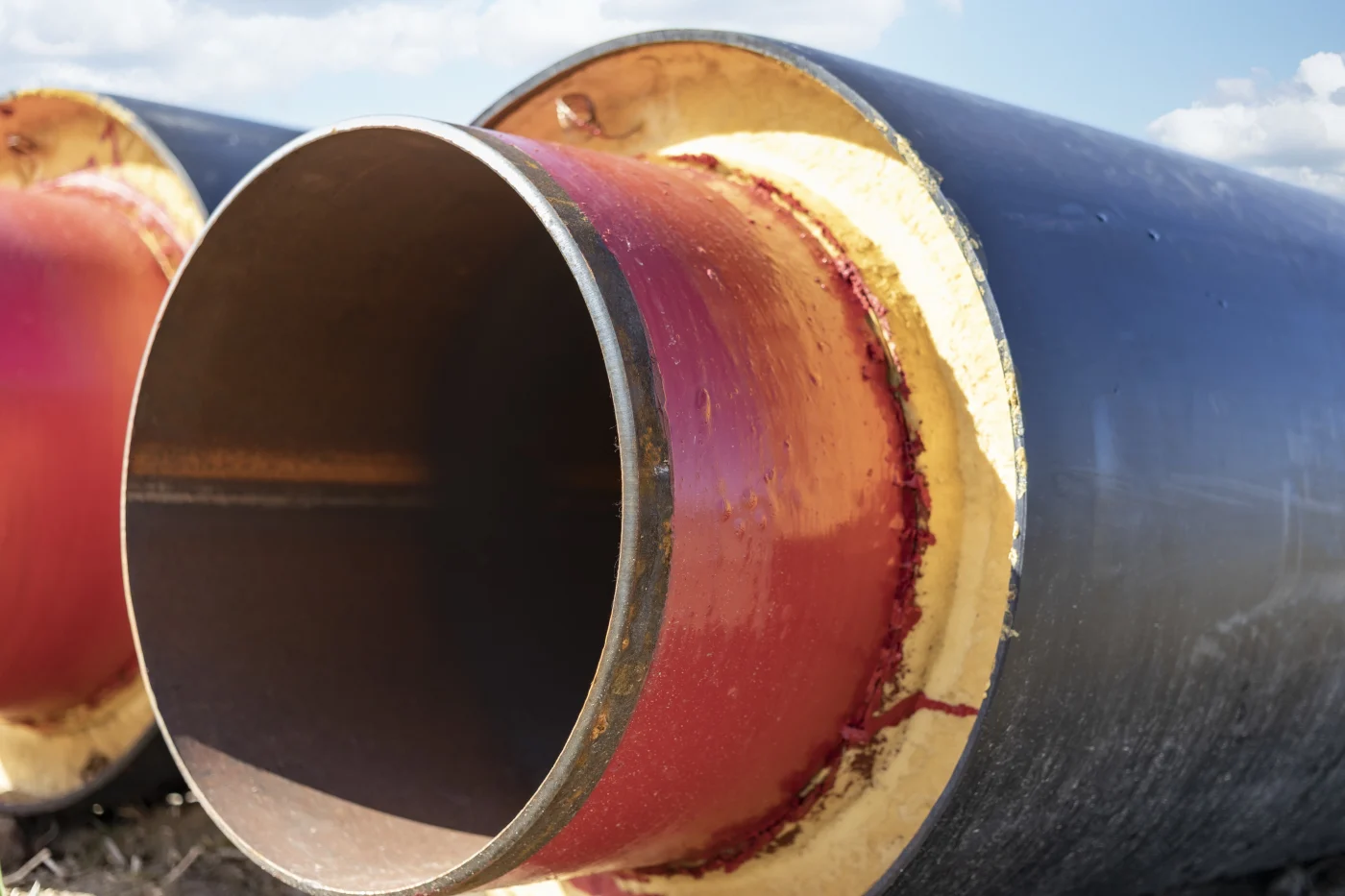 Close-up of large sewer pipe with red liner, yellow insulation, black outer casing, open end, metal rim, and cloudy sky beyond.