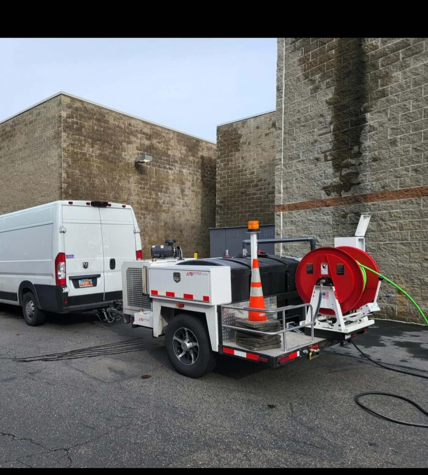 Hydro jetting trailer with red hose reel, white van, orange cone, green hose, and wet pavement beside a stained brick commercial wall.