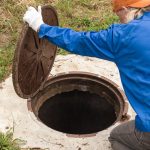 A worker opens a sewer manhole cover beside grass, wearing a blue jacket, an orange cap, and white gloves near a concrete access opening.