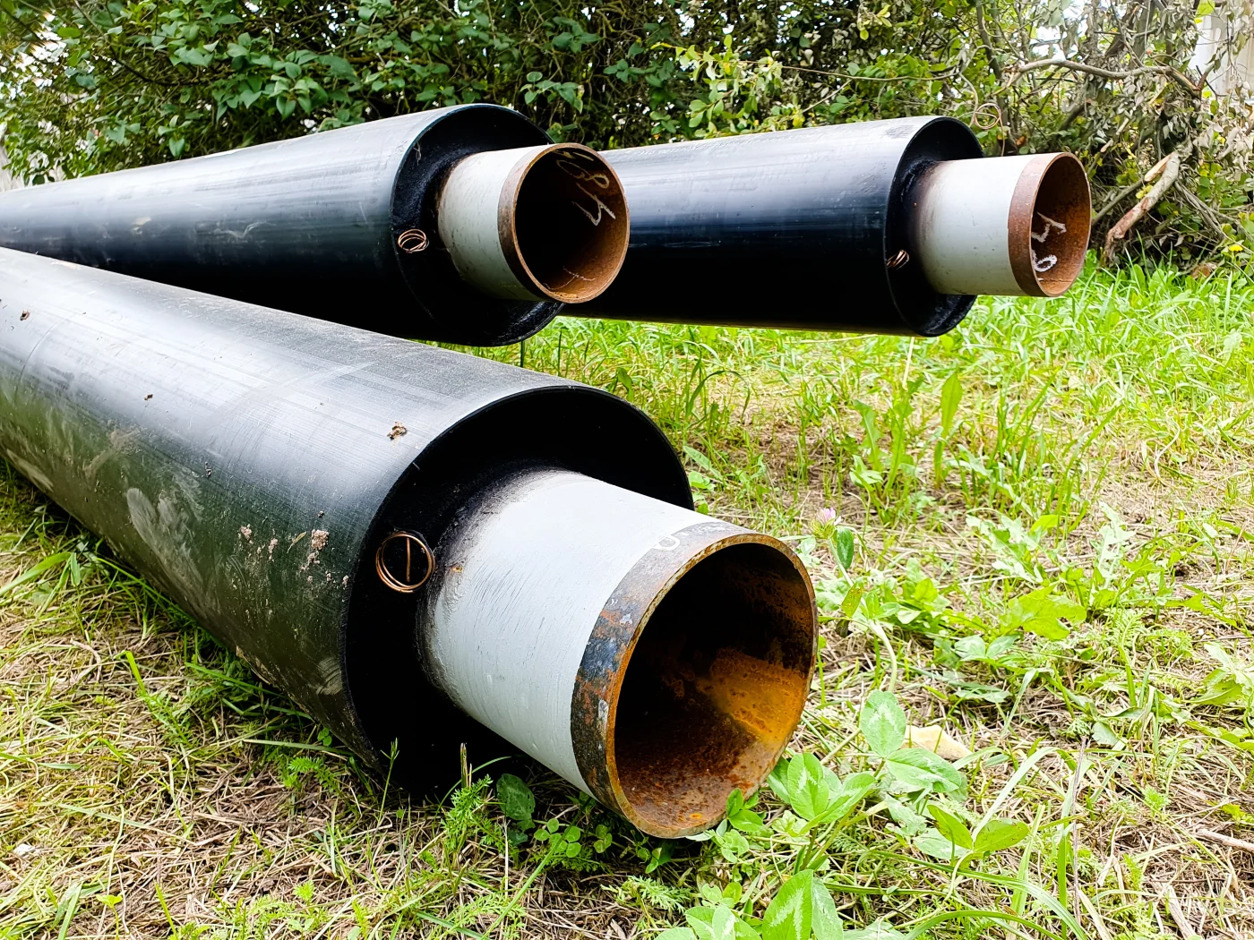 Black commercial sewer pipes with gray inner liners rest on grass, showing open pipe ends, metal rings, rusted edges, and greenery nearby.